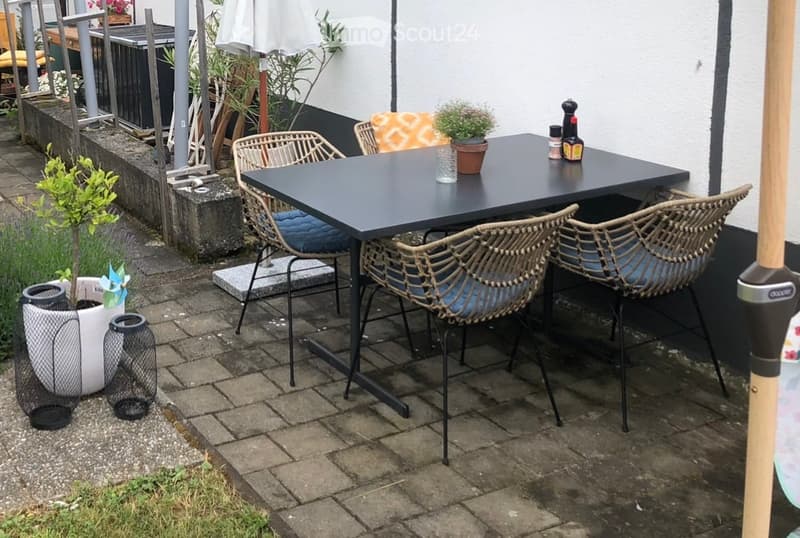 Outdoor patio area with a black rectangular table, four wicker chairs with blue cushions, potted plants, and decorative lanterns.