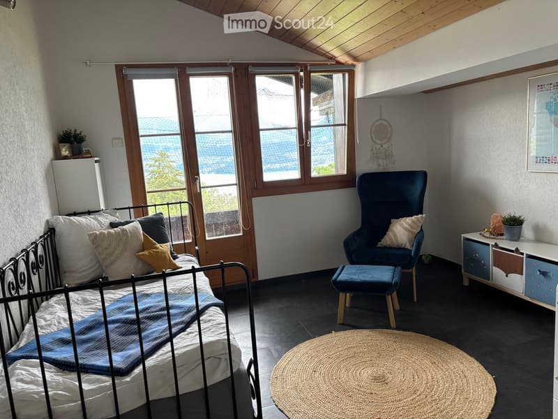 Cozy bedroom with a metal-framed bed, a blue armchair, a round woven rug, a small table with drawers, and a dreamcatcher on the wall. Large windows offer a scenic view.