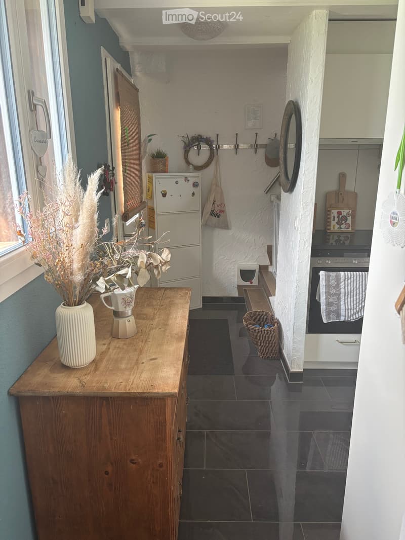 The kitchen features a wooden table with a vase and cup, a refrigerator, an oven, a cutting board, a basket, a mirror, and a wreath on the wall.