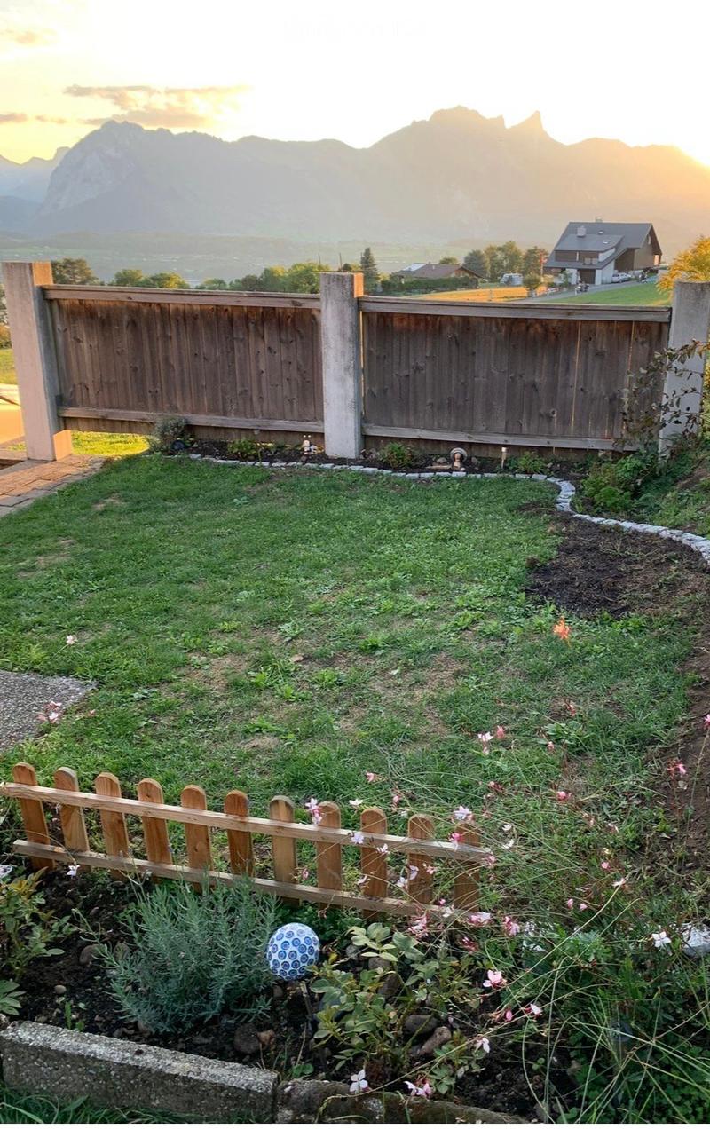 A well-maintained garden with a wooden fence, green lawn, and flower beds. The background features a scenic mountain view and a neighboring house.