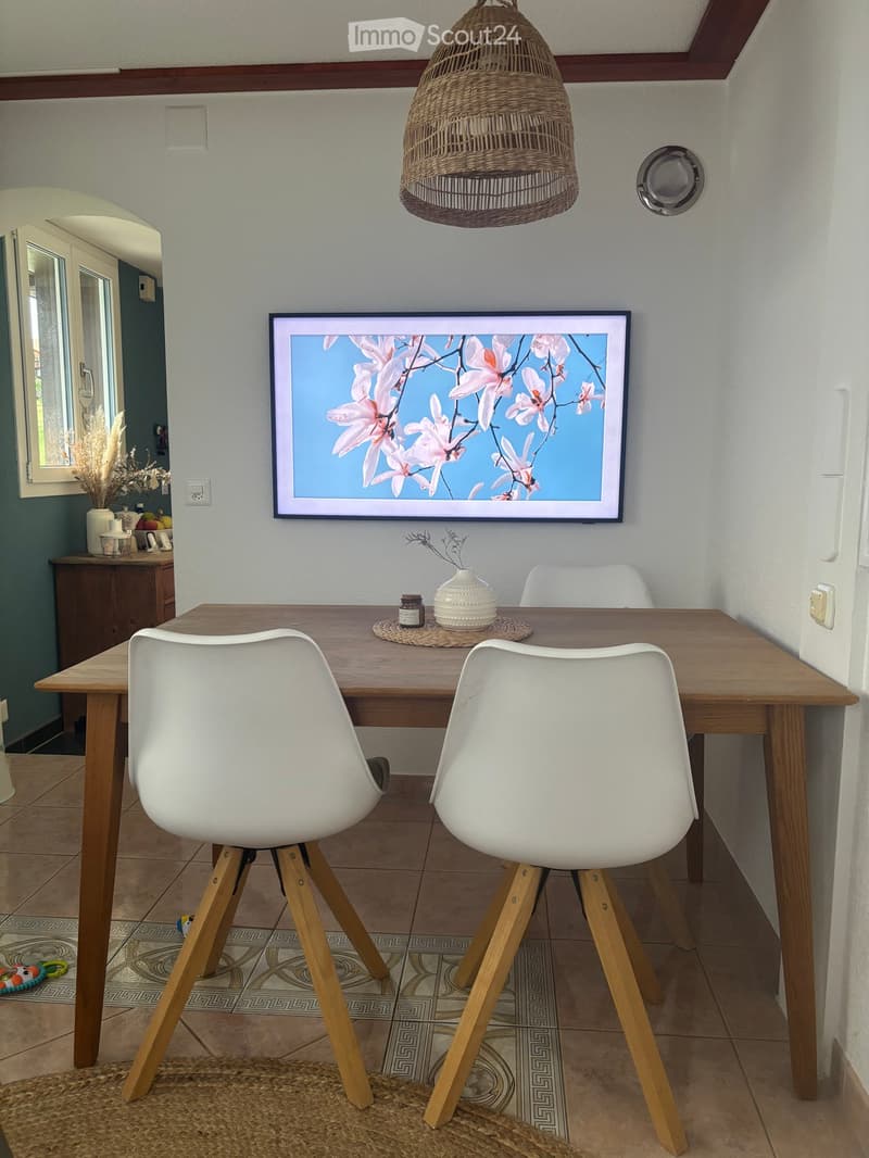 Dining area with a wooden table, two white chairs, a hanging woven pendant light, a mounted screen displaying a floral image, and a small cabinet with decorative items.