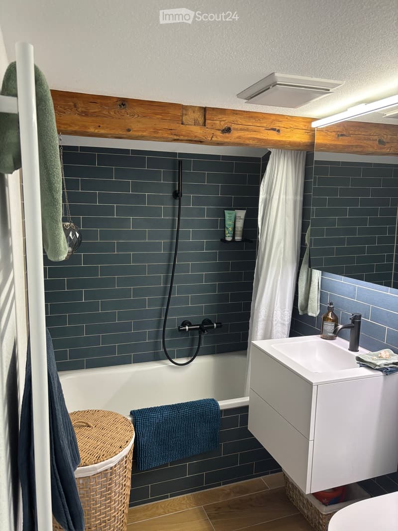 Bathroom with a bathtub, sink, and modern fixtures. Features dark blue tiles, a wooden shelf, and a wicker basket.