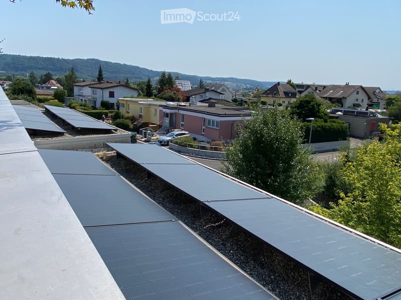Roof with solar panels, gravel surface, surrounding houses, greenery, and distant hills.