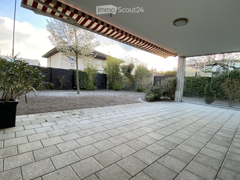 Paved patio area with a striped awning, surrounded by greenery and a wooden fence.