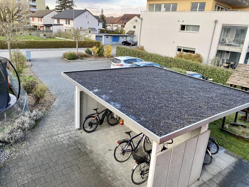 Modern residential building with a covered parking area, bicycles parked underneath, and a gravel-covered rooftop terrace.
