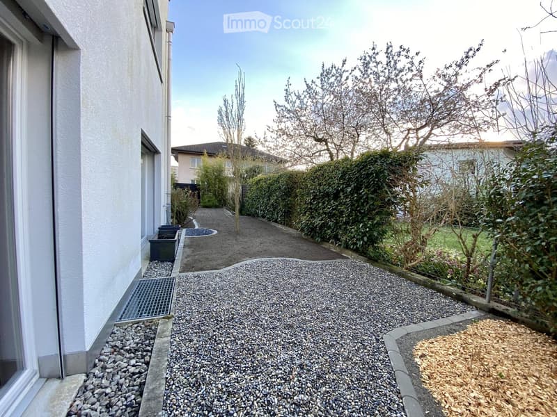 Gravel pathway leading to a house, surrounded by greenery and shrubs, with a small garden bed on the right.
