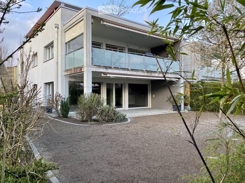 Modern two-story house with large glass windows and a balcony on the upper level, surrounded by greenery and a gravel driveway.