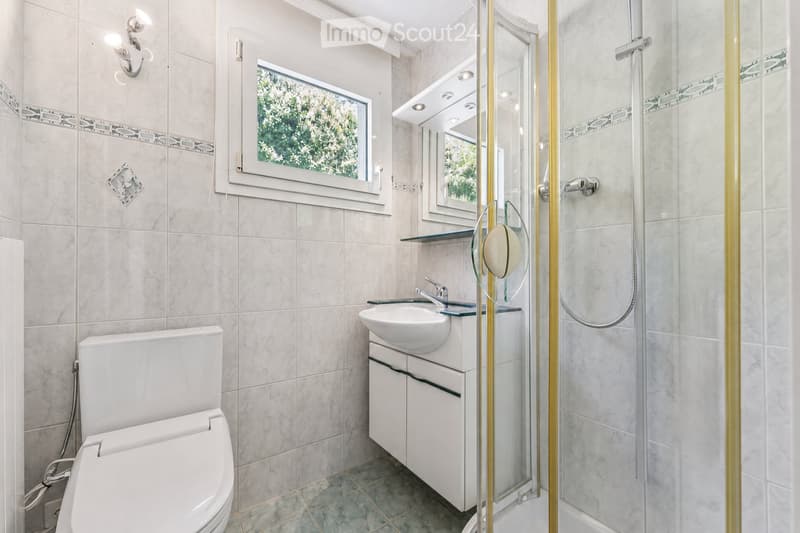 Modern bathroom with white tiles, a toilet, a sink with a cabinet, a glass shower enclosure, and a mirror above the sink. The room features a window with a view of greenery.