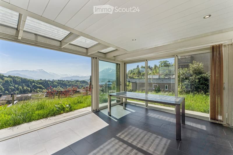 Bright, modern room with large glass walls and a retractable roof, offering panoramic mountain views. Features a sleek, minimalist table in the center.