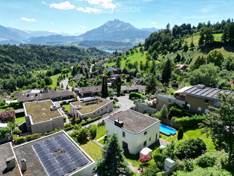 Aerial view of a residential area with modern houses, green lawns, a swimming pool, and a scenic mountain backdrop.