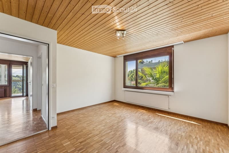 Empty room with wooden flooring, white walls, and a large window with a view of greenery. The ceiling is wooden, and there is a radiator below the window.