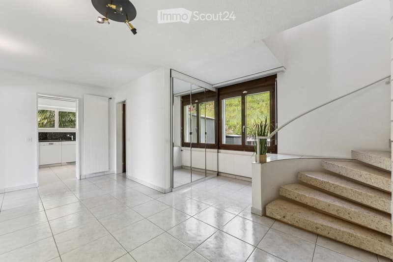Bright, modern hallway with white walls, tiled floor, large windows, and a staircase leading upwards. Features a sleek, contemporary design with a small plant on a pedestal near the stairs.