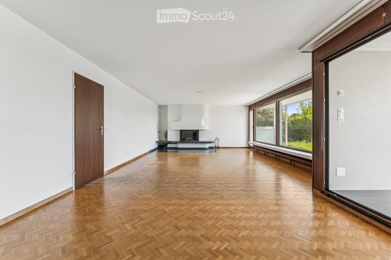 An empty room with wooden herringbone flooring, white walls, large windows, and a fireplace area in the background.