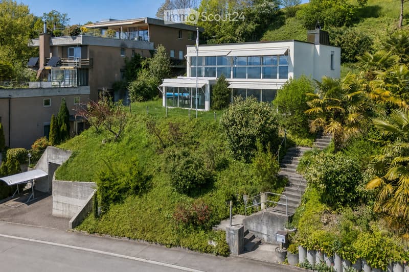 A modern two-story house with large windows and a balcony, surrounded by lush greenery and trees on a hillside.
