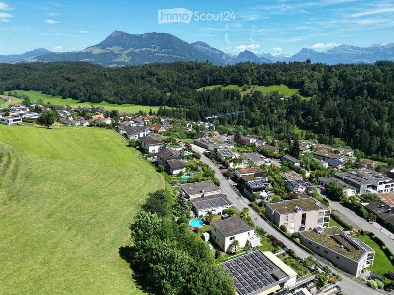 Aerial view of a suburban neighborhood with multiple houses, green lawns, trees, and a swimming pool visible in the foreground, surrounded by forested hills and mountains.