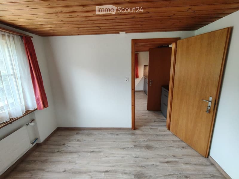 Unfurnished Room. Empty hallway with wooden flooring, white walls, and a wooden door leading to another room. There is a window with a red curtain on the left side.