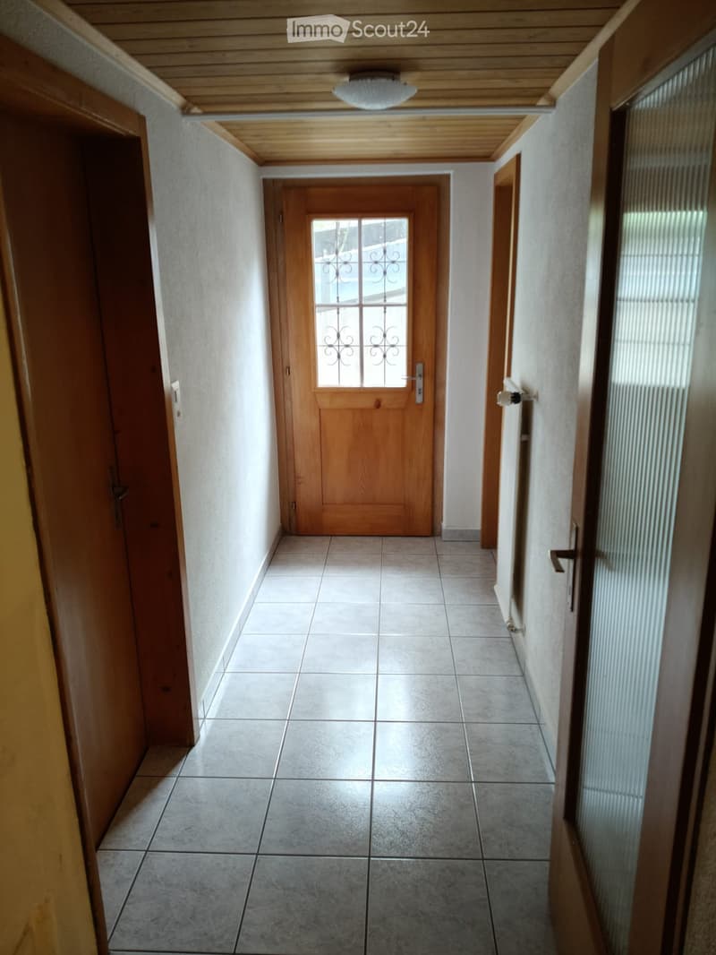 Corridor / Hallway. A clean, tiled hallway with wooden doors and a central entrance door featuring a decorative glass panel. The ceiling has wooden paneling and a recessed light fixture.