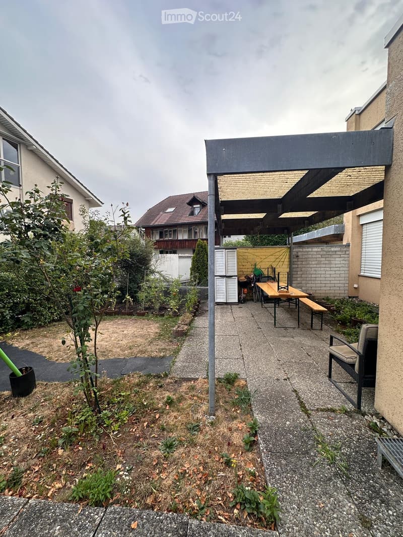 Garden area with a patio, table, chairs, and a bench under a covered structure. There are plants and a small garden bed on the left side.