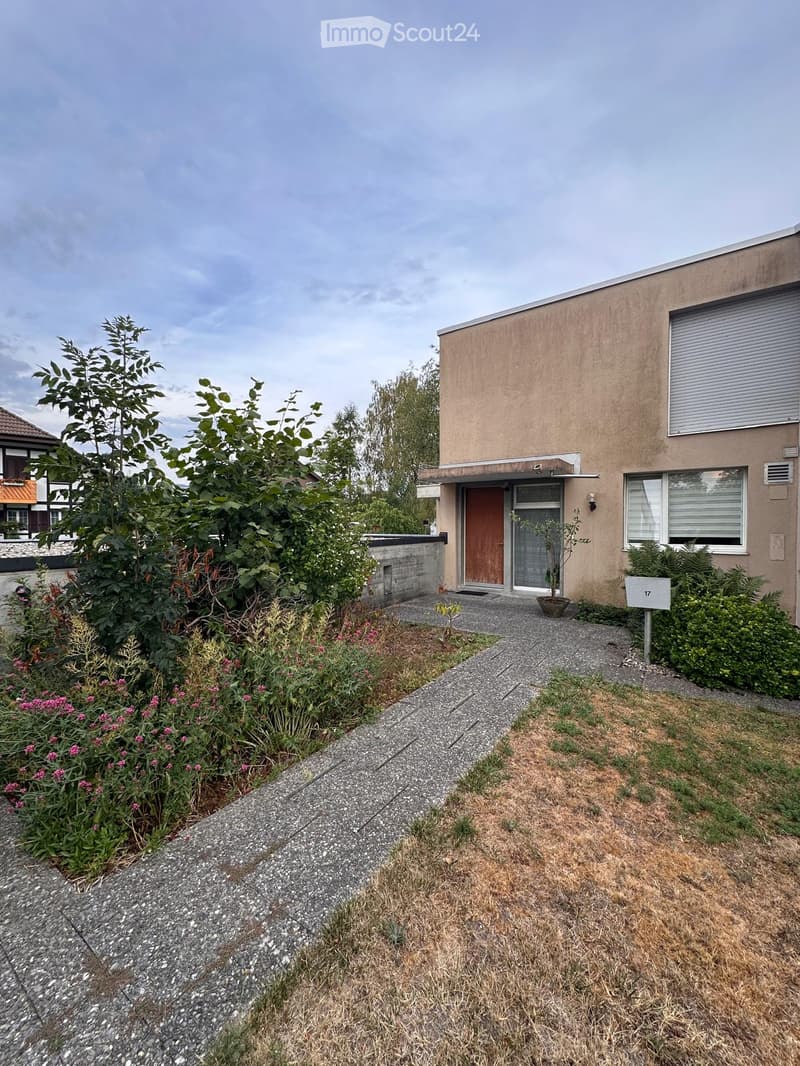 Single-story house with a beige facade, a wooden front door, and a small garden area with various plants and flowers.
