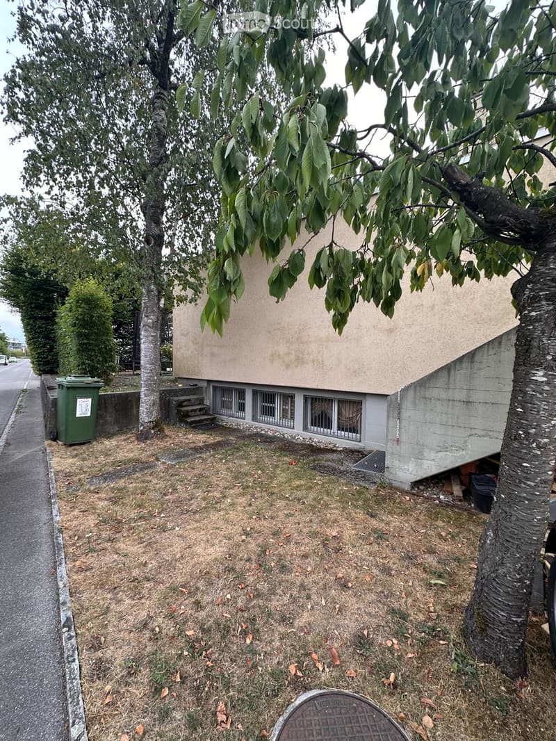 A small, modern building with a beige facade and grey concrete elements, surrounded by trees and a grassy area.