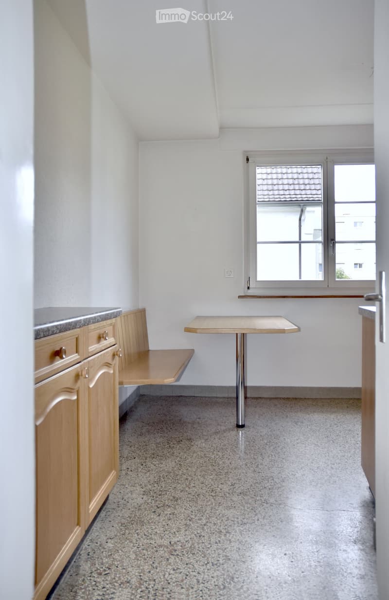 A small, furnished room with a table, bench, and cabinet, featuring a window with a view of a roof and a door.