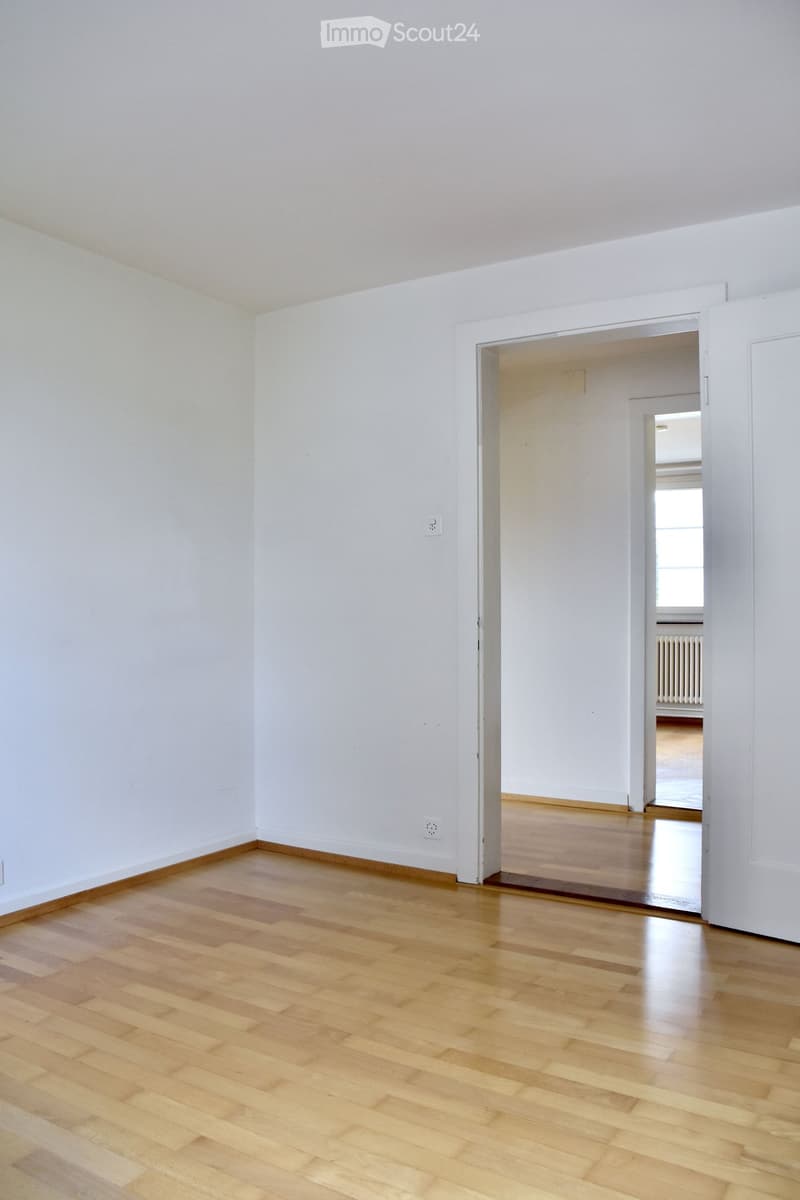Empty room with light wooden flooring and white walls, featuring an open doorway leading to another room with similar flooring and white walls.
