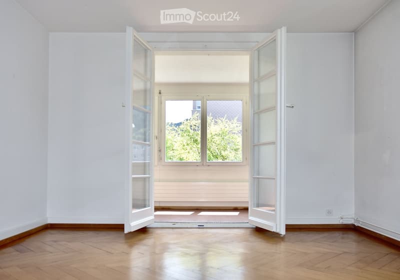 Empty room with wooden flooring, white walls, and large windows with open doors leading to another space.