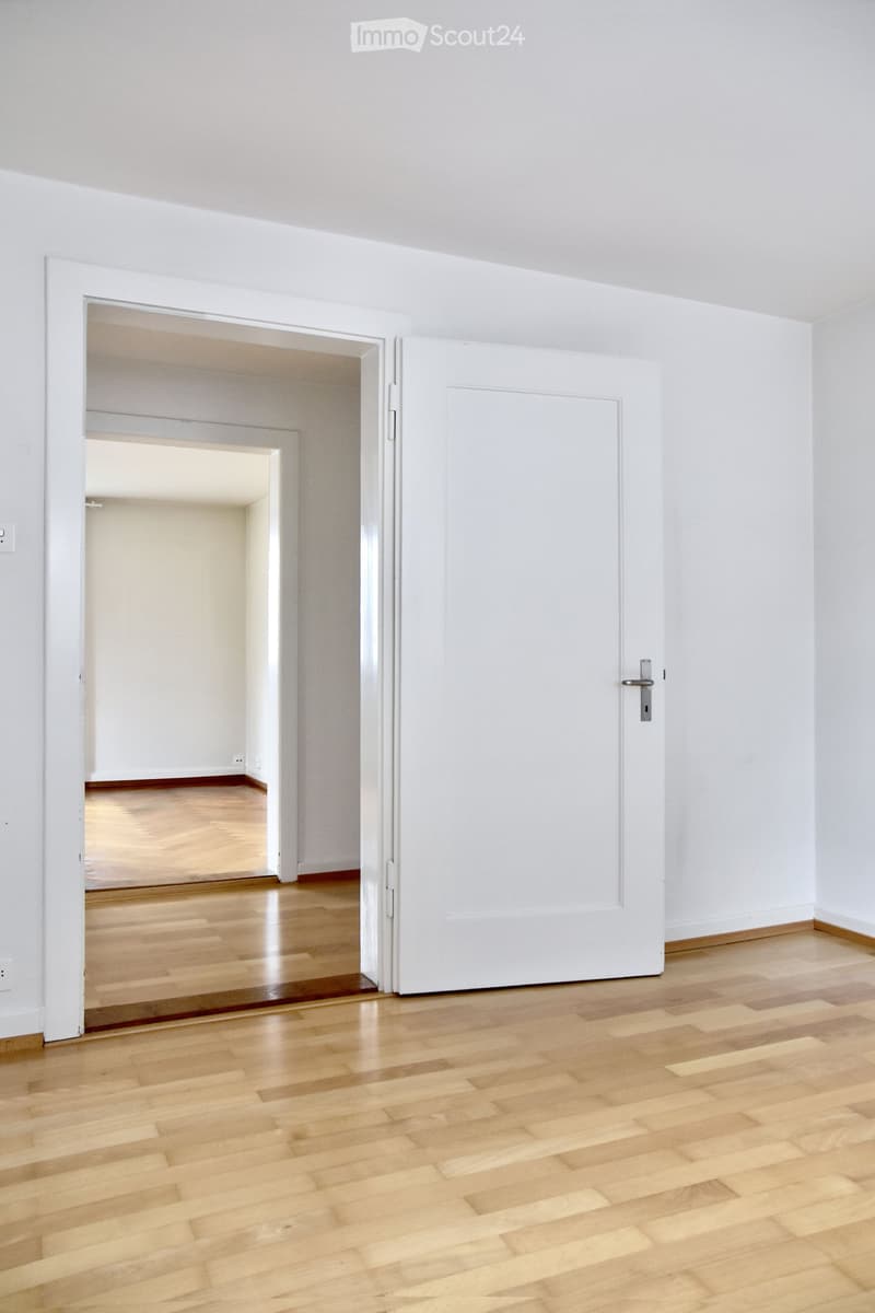 Empty room with light wooden flooring and white walls, featuring an open doorway leading to another similarly furnished space.