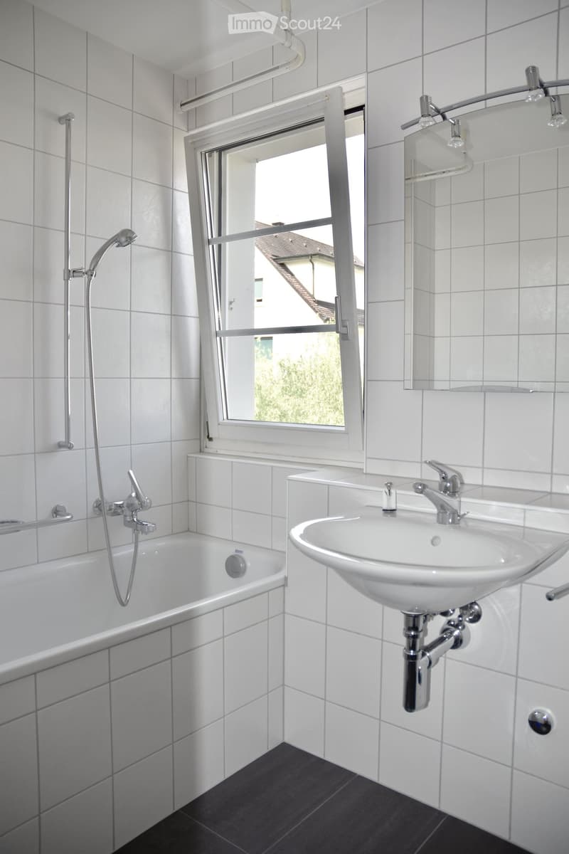 Modern bathroom with white tiled walls, a bathtub, a sink with a chrome faucet, and a large window offering a view of greenery outside.