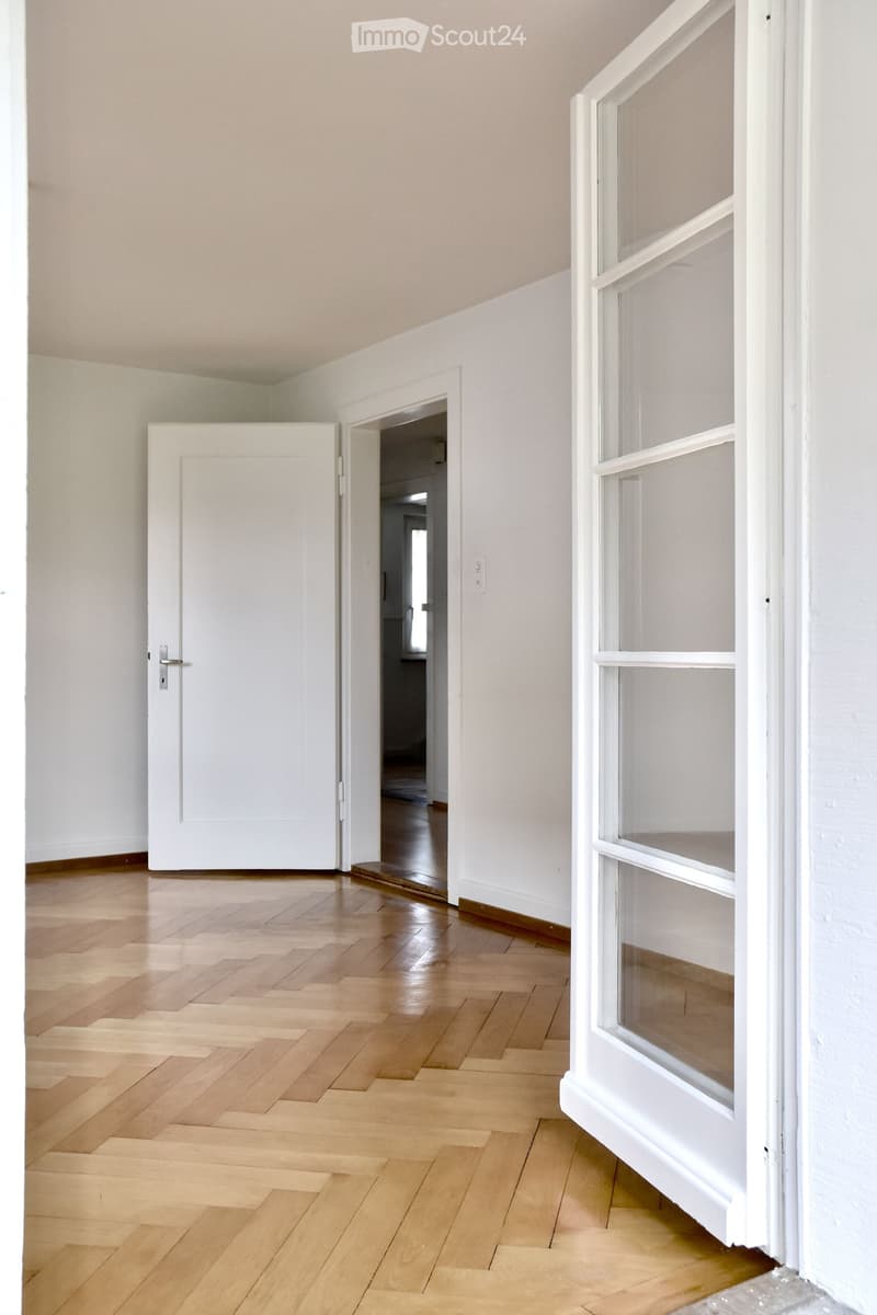 Empty hallway with wooden flooring, white walls, and a large window on the right side. A white door is open, leading to another room.