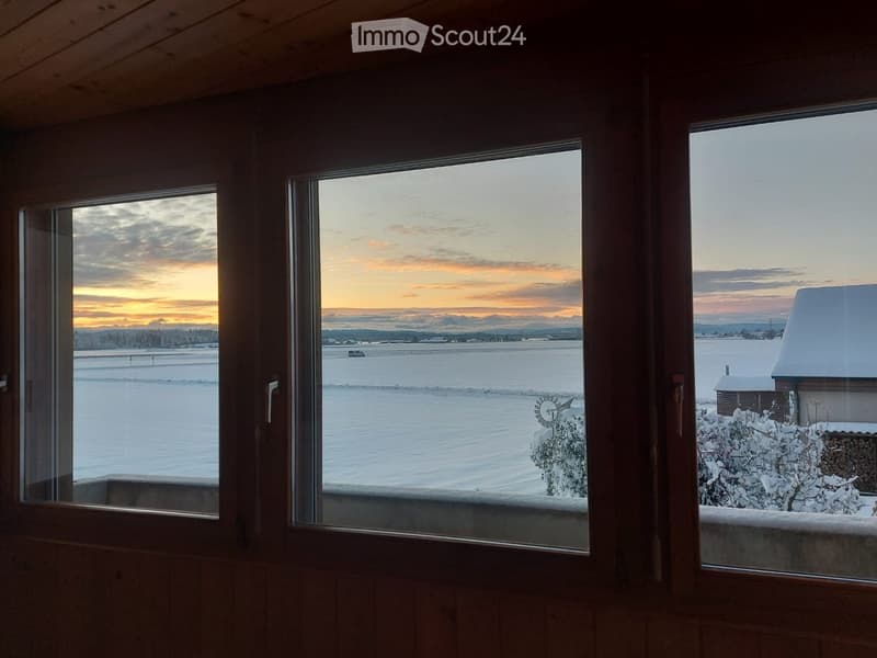 Wooden-framed balcony with large windows overlooking a snowy landscape at sunset, featuring distant mountains and a small cabin.
