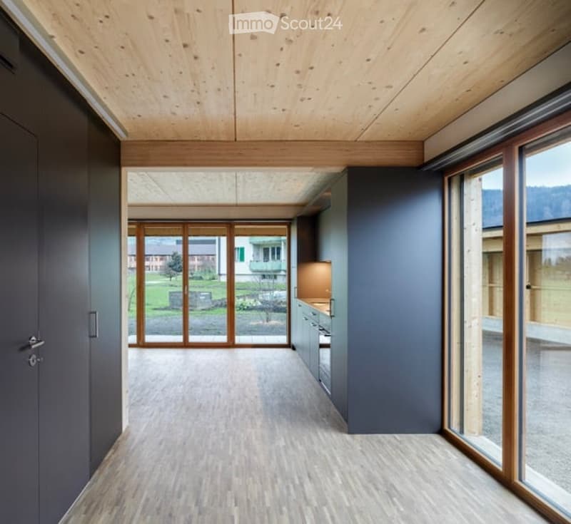 Open-plan unfurnished space with wooden ceiling and floor, large glass doors leading to outdoor view, grey walls, and minimal built-in cabinetry.