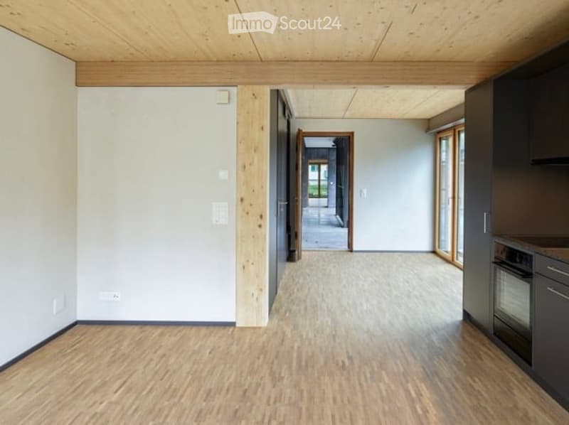 Empty room with wooden flooring, white walls, and a wooden ceiling. There is a doorway leading to another room and a kitchen area visible on the right side.