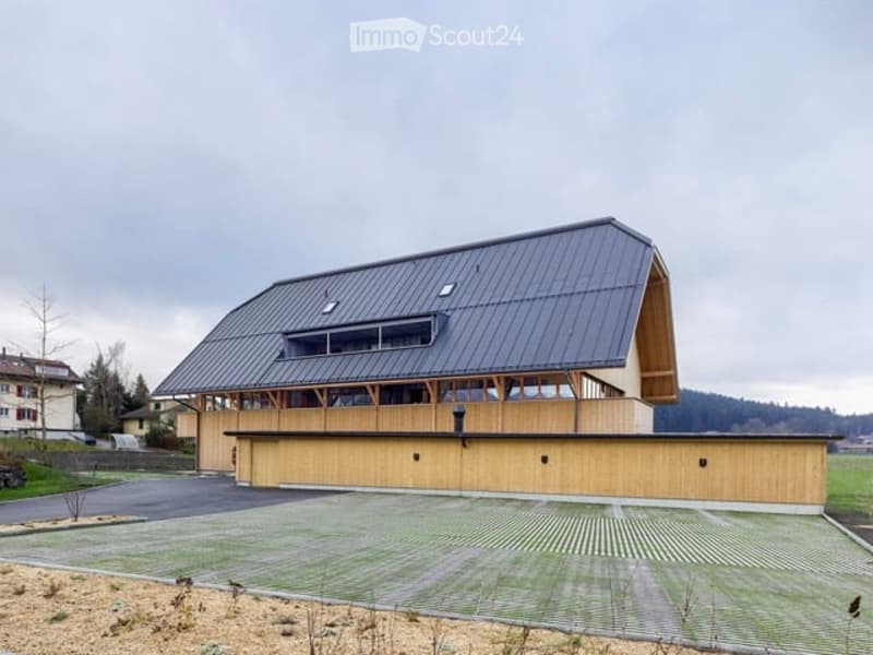 Modern two-story building with a large garage, wooden facade, and metal roof. The structure features large windows and a spacious driveway.