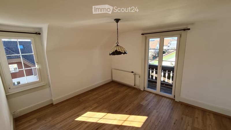 Empty room with wooden flooring, white walls, large window, sliding glass door leading to a balcony, and a hanging light fixture.