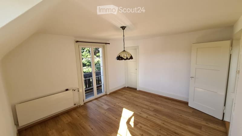 Empty attic room with wooden flooring, white walls, a hanging light fixture, and a large window with a view of the garden.