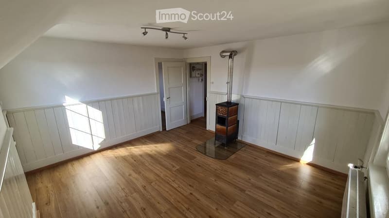 Empty room with wooden flooring, white walls, and a fireplace in the corner. Natural light streams in from a window on the left side.