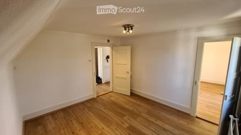 Empty room with wooden flooring, white walls, and a ceiling light fixture. There is an open doorway leading to another room.