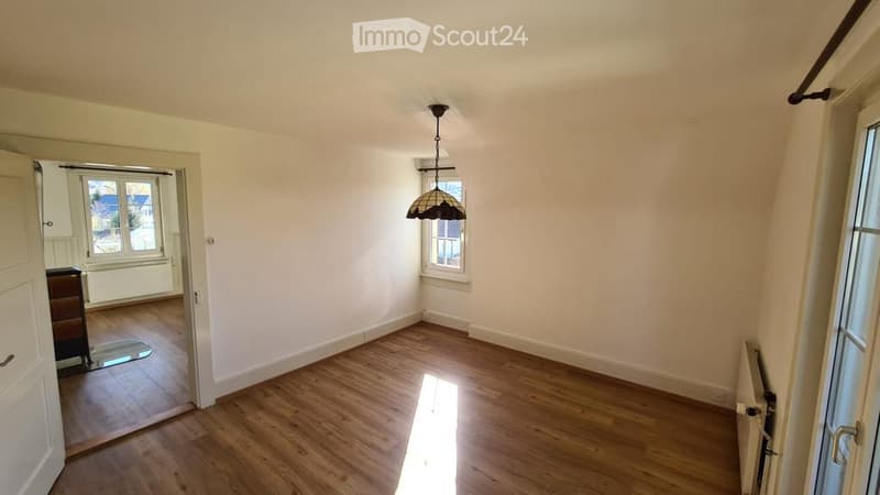 Empty room with wooden flooring, white walls, and a hanging light fixture. There is a doorway leading to another room with a window and radiator visible.