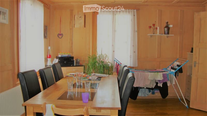 A well-lit dining area with a wooden table surrounded by black chairs, a potted plant, and a shelf with decorative items. The room features wooden walls and a drying rack with clothes in the background.