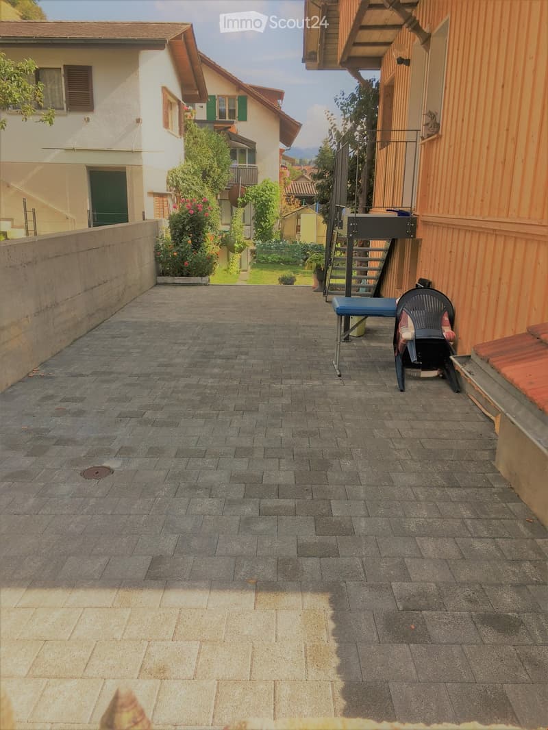 Paved outdoor area with a small table and chair, surrounded by buildings and greenery.