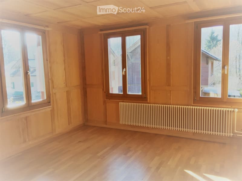Empty room with wooden flooring, wooden walls, large windows, and a radiator.