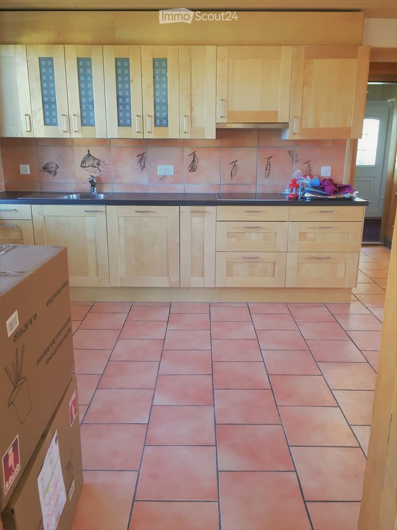 Empty kitchen with light wooden cabinets, black countertop, and terracotta tiled floor. A sink and faucet are visible, with a few items on the counter.
