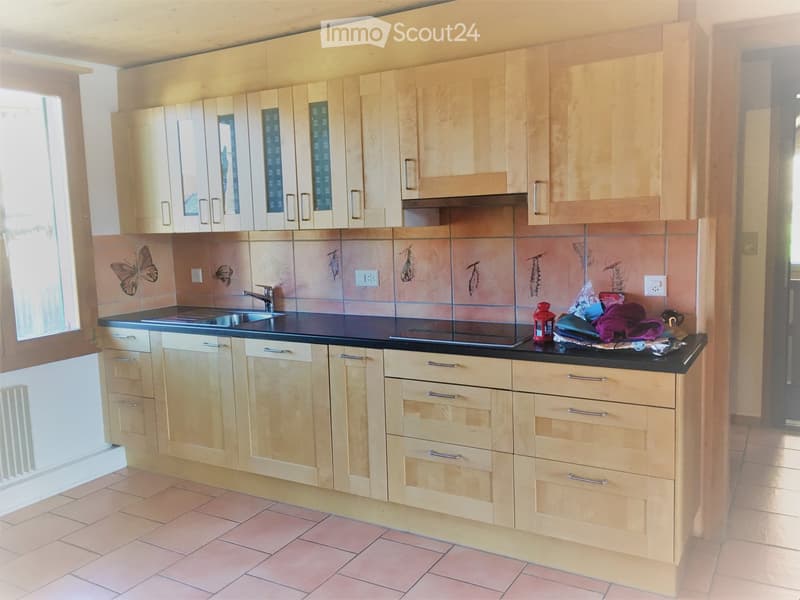 Modern kitchen with light wooden cabinets, black countertop, sink, stove, and various small items on the counter.