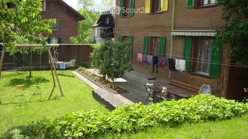 A well-maintained garden with a swing set, clothesline, patio furniture, and surrounding houses.