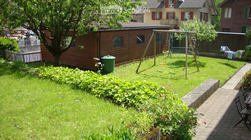 Well-maintained garden with green grass, a swing set, a wooden shed, a tree, and a clothes drying rack.