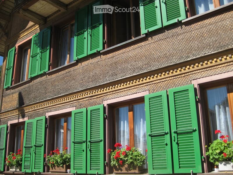 Traditional wooden building with green shutters, multiple windows, and flower boxes with red flowers on the balconies.