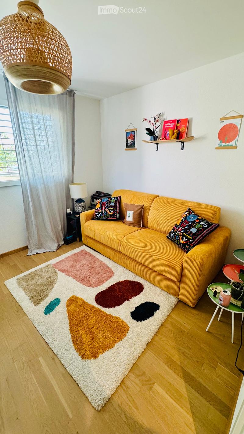A modern living room with a mustard yellow sofa, colorful pillows, a large geometric rug, a small side table with a lamp, and shelves with books and decorative items.