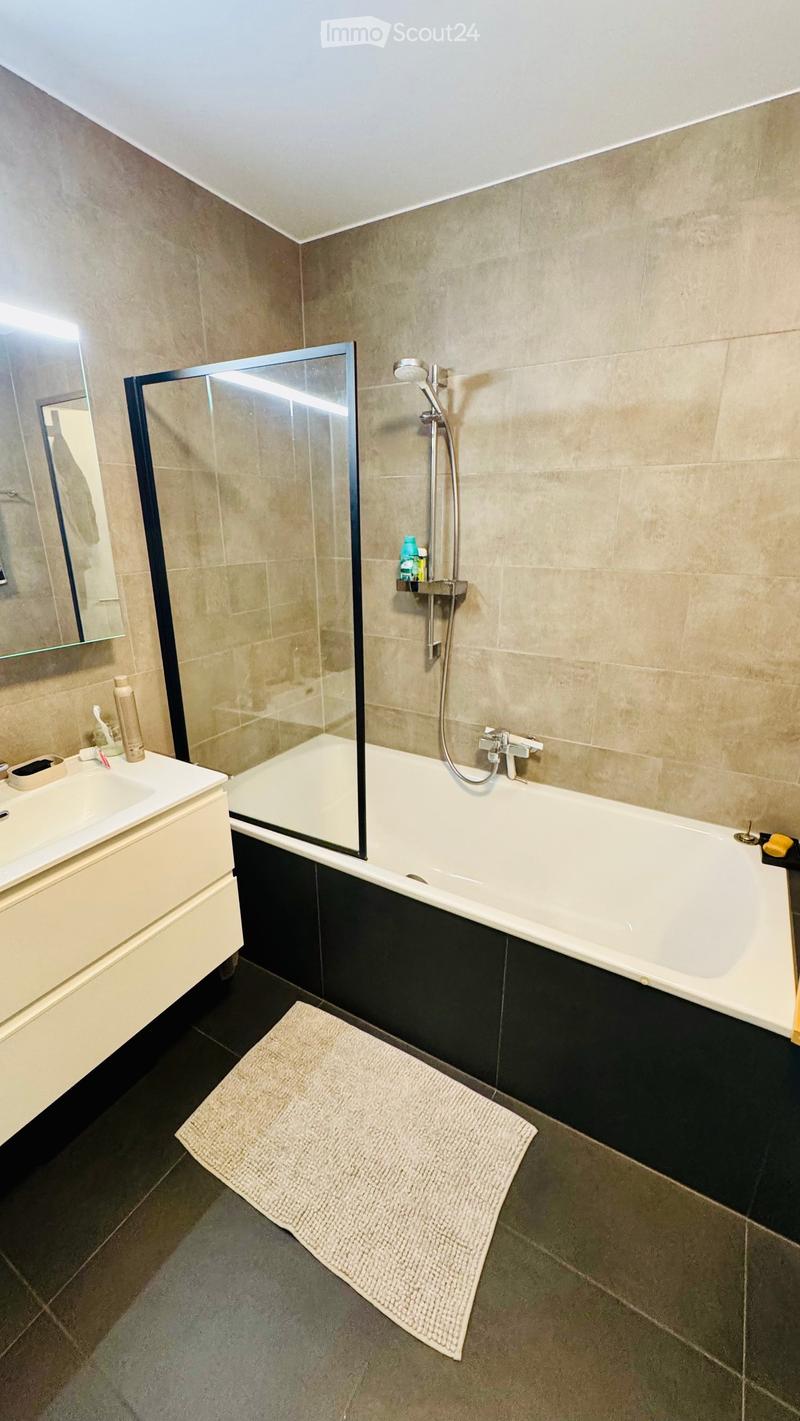 Modern bathroom with a bathtub, shower, sink, and mirror. Features a white bathtub with a glass shower enclosure, a white sink with a cabinet, and a beige bathroom rug on the floor.