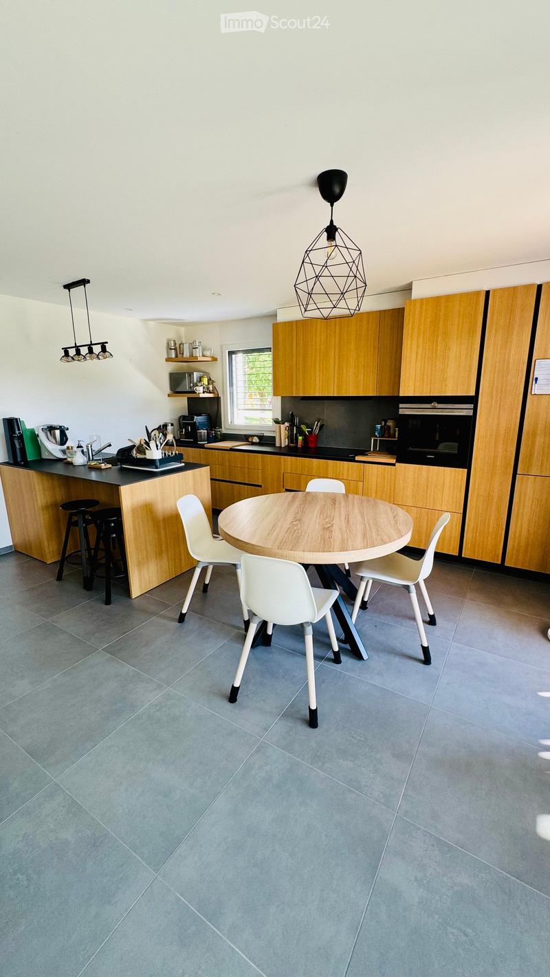 Modern kitchen with light wooden cabinets, a round dining table with four white chairs, and a bar counter with stools. Appliances include a microwave, oven, and blender on the counter.